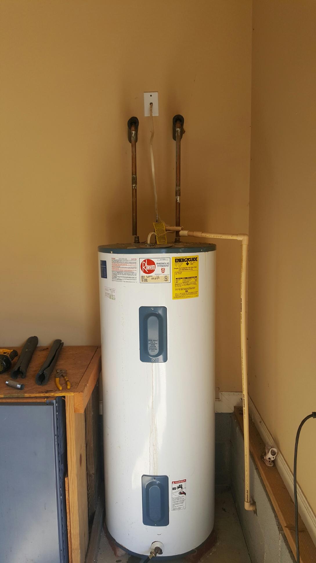 A white water heater is sitting in a corner of a room next to a wooden table.