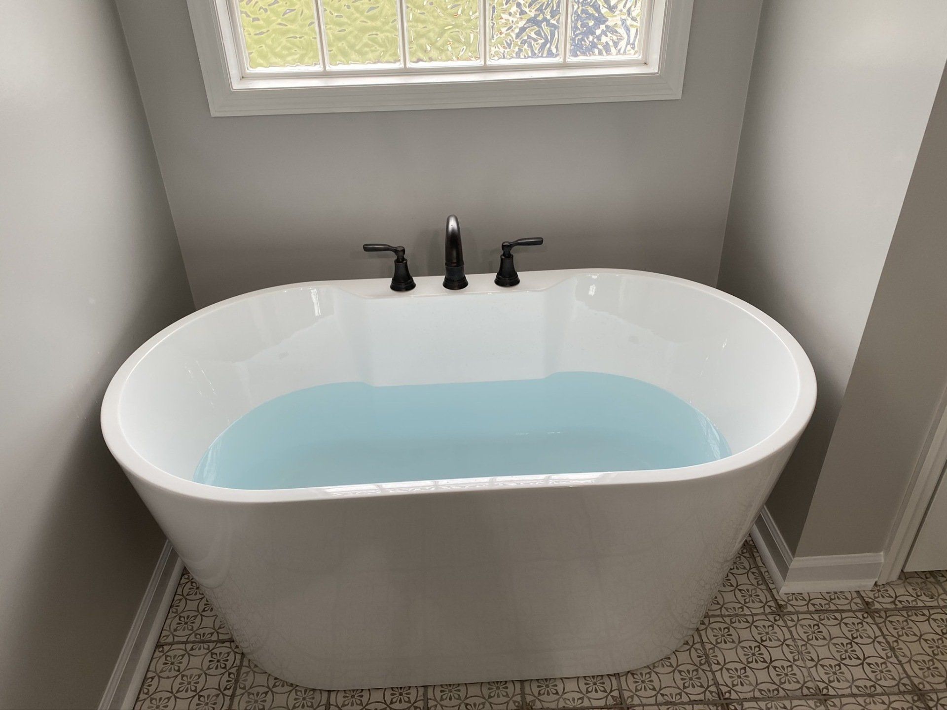 A white bathtub is filled with blue water in a bathroom next to a window.