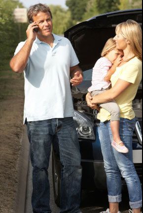 A man is talking on a cell phone while a woman holds a child in front of a broken down car.