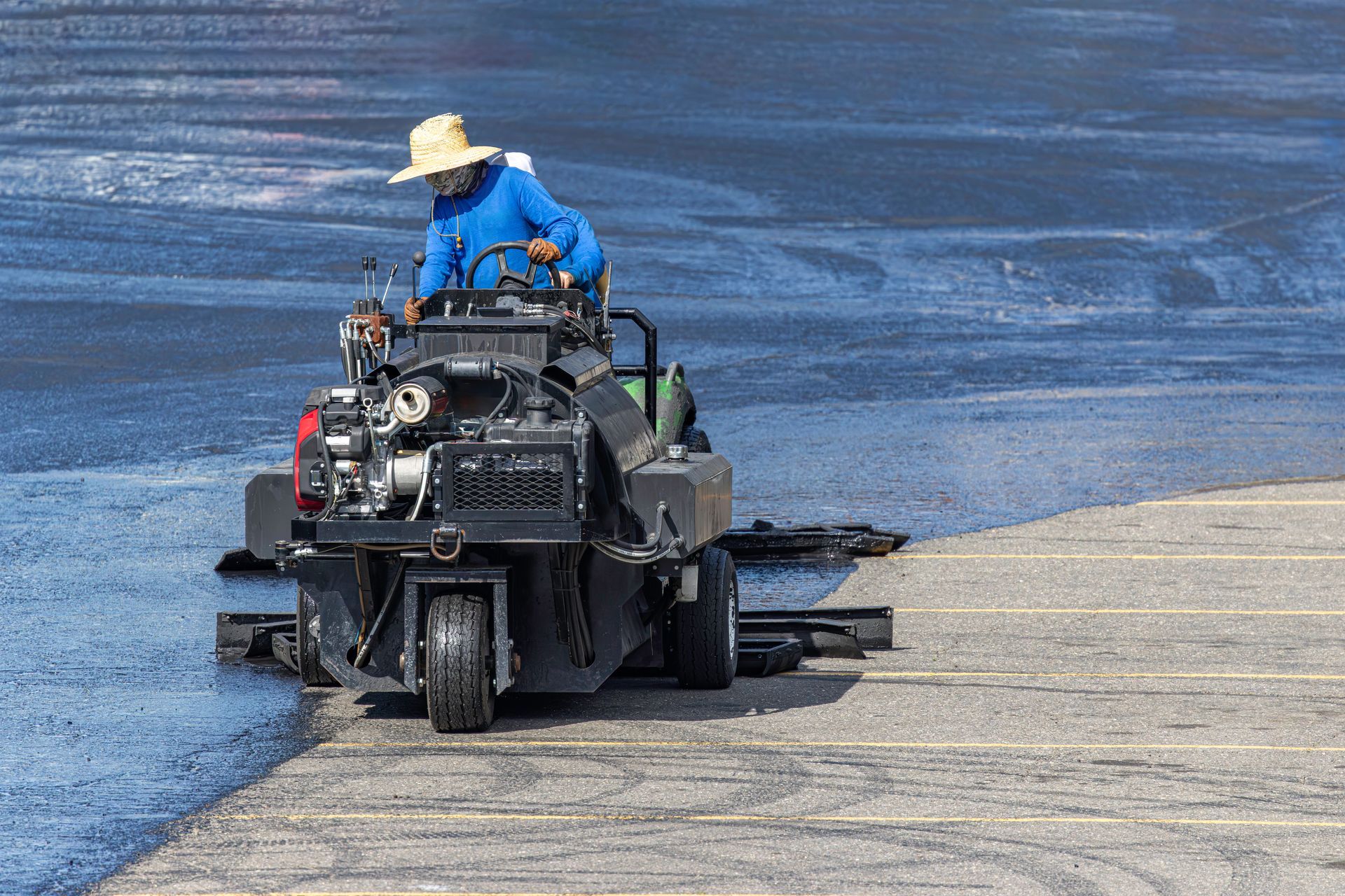 Worker operating a ride-on sealcoating machine spreading black sealer across a striped parking lot.