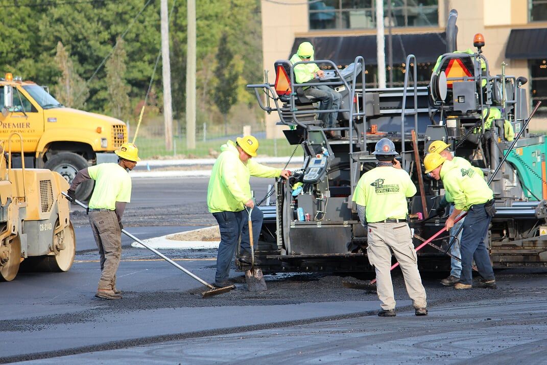 Road construction crew paving asphalt on a sunny day, wearing yellow safety vests and hard hats.