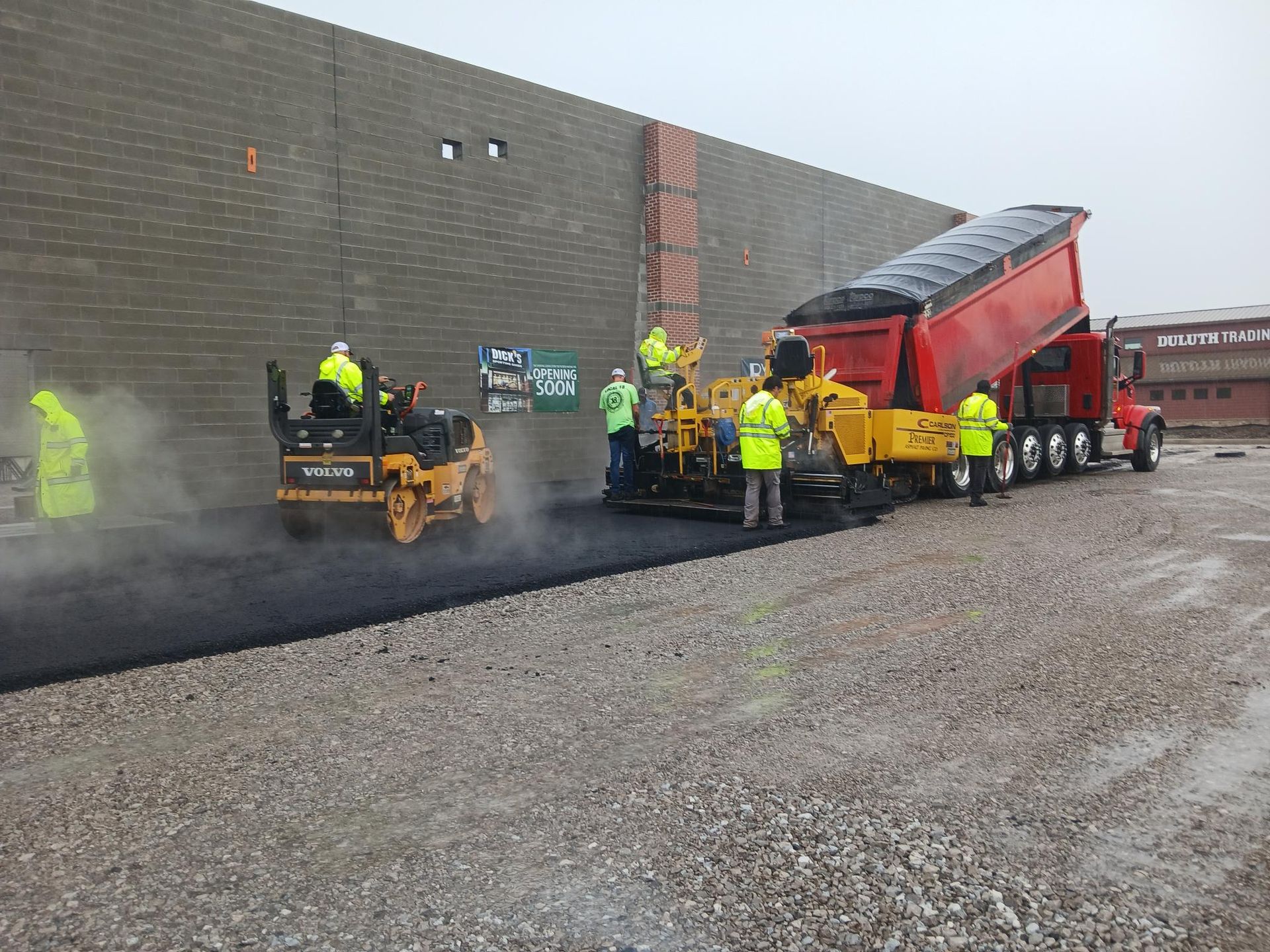 Workers paving asphalt beside a red dump truck and steamroller near a gray wall
