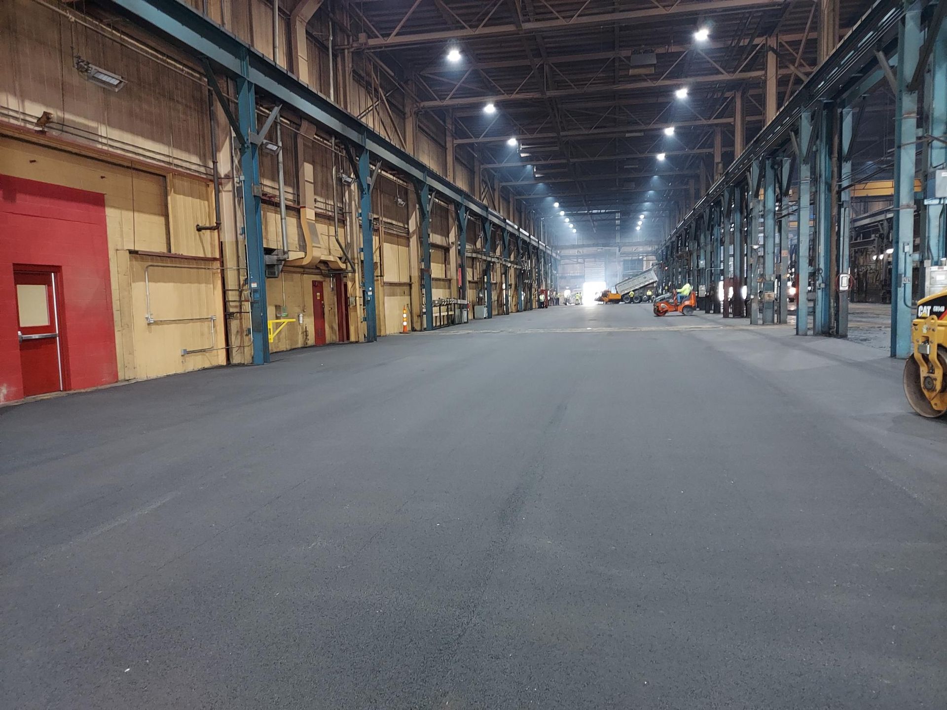 Empty warehouse interior with a wide concrete aisle and rows of steel beams, lit by overhead lights