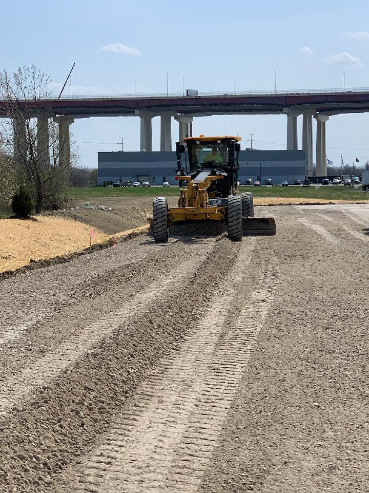 A yellow motor grader smoothing a gravel road with bridge and industrial buildings in the background.