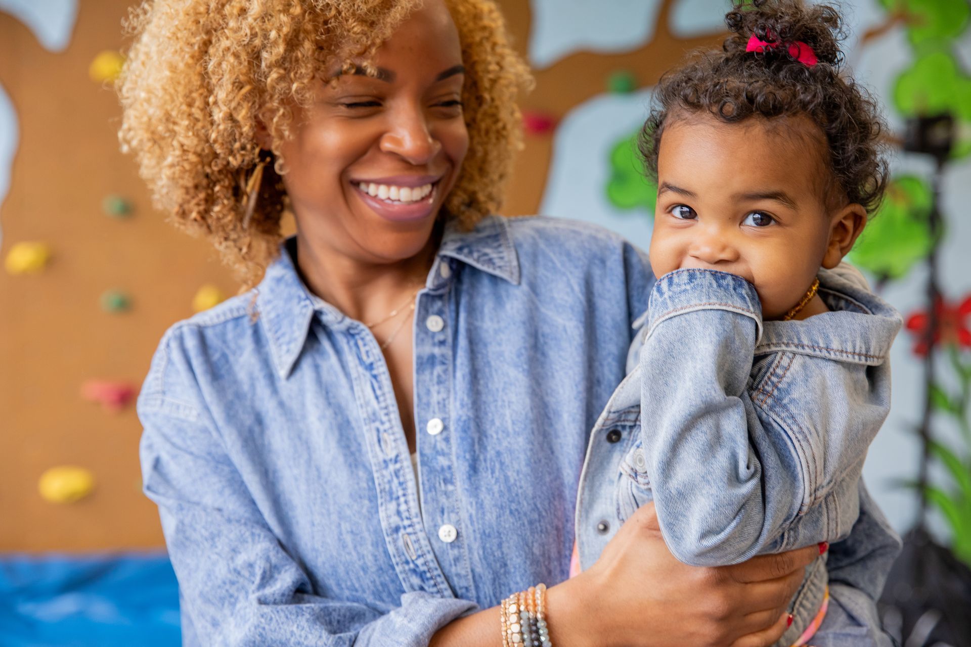 Woman holding a baby; both smiling. The woman wears denim, the baby a denim jacket. Indoors, playroom setting.