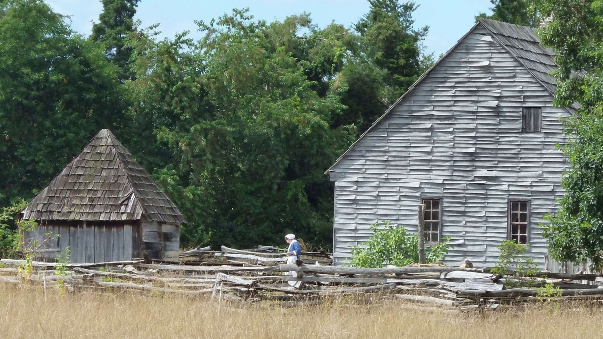 Old, weathered wooden buildings behind a split-rail fence; a person walks along a path.