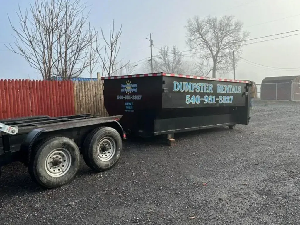 Black dumpster on a trailer, ready for transport. Exterior setting with a red fence and bare trees.