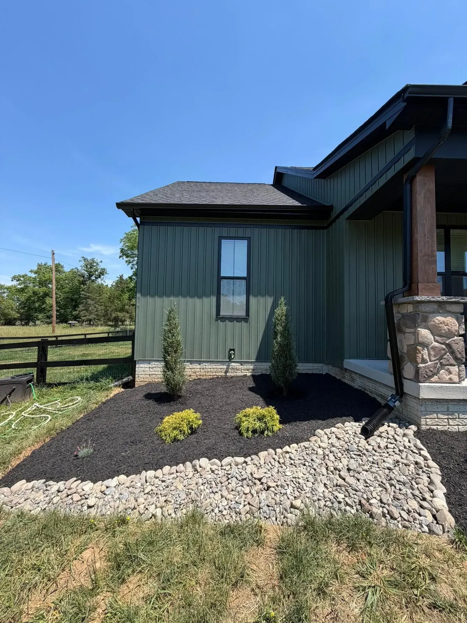 Front of a house with dark green siding, stone entry, and small landscaped shrubs under a blue sky.