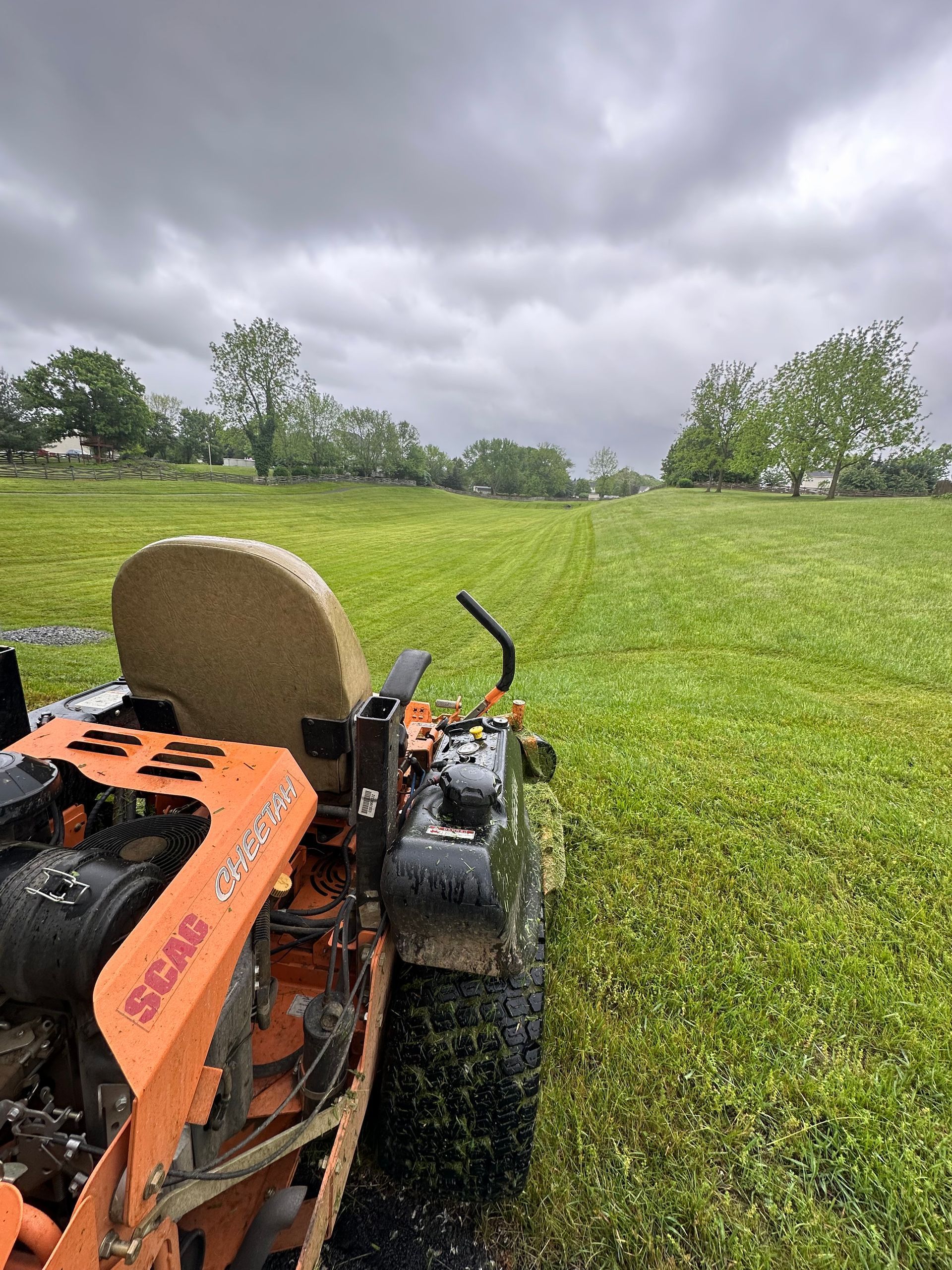 Orange lawnmower cutting grass in a large field on an overcast day.