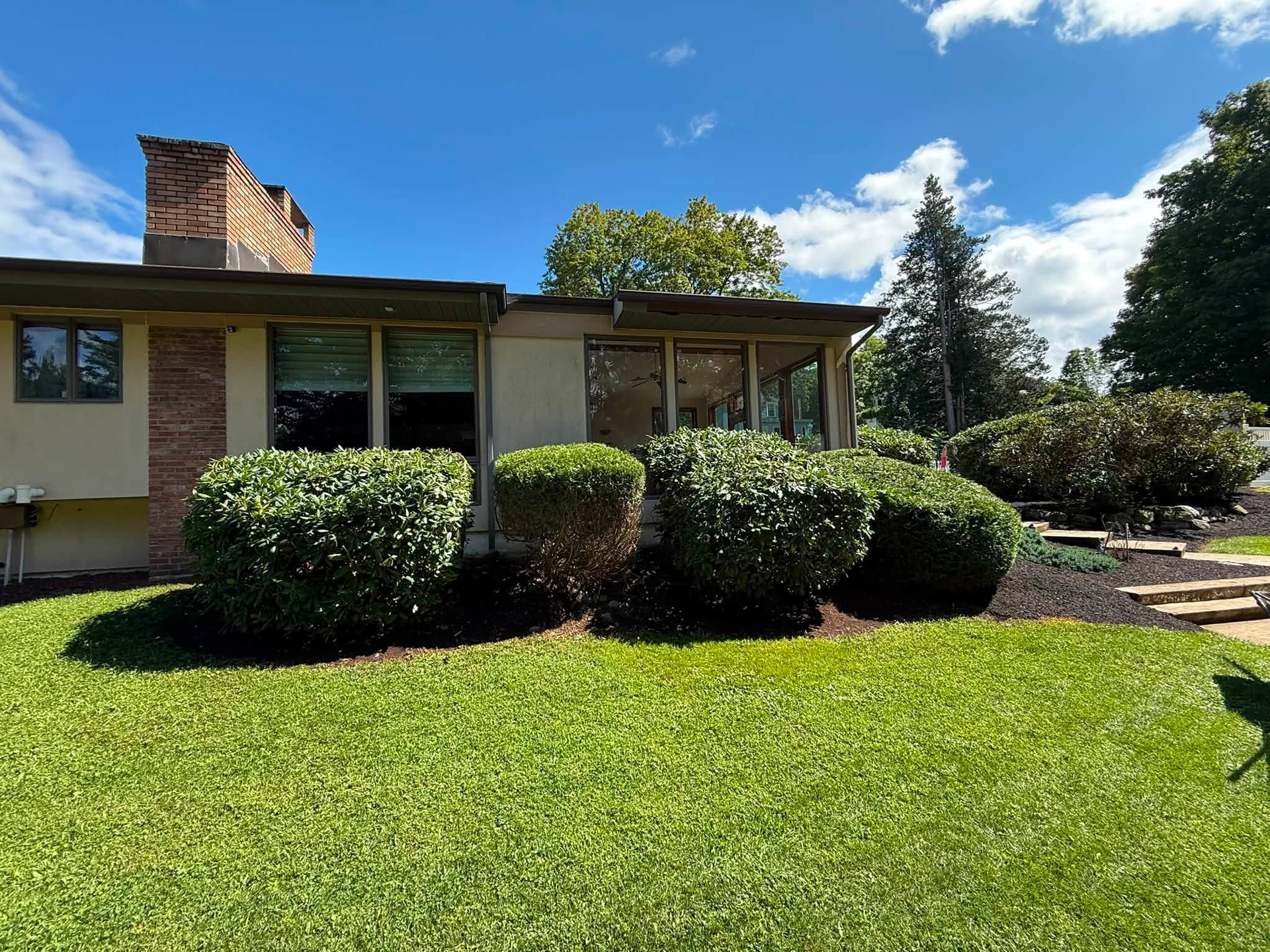 Single-story house with brick chimney, trimmed bushes, and a bright green lawn under a blue sky