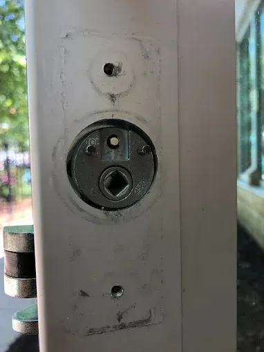 Close-up of a white door with a circular deadbolt mechanism exposed, featuring a central square hole and mounting holes.