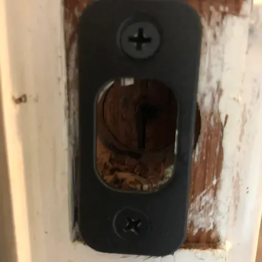 A black rectangular strike plate is screwed into a door frame, showing an empty, hollow opening leading to raw wood.