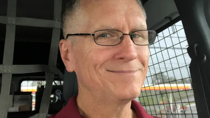 A person with glasses smiles while sitting in the driver's seat of a vehicle with a cargo partition visible behind them.