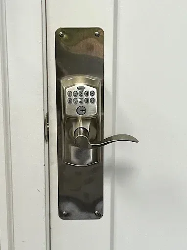 A silver electronic keypad door handle with a metal mounting plate installed on a white door.