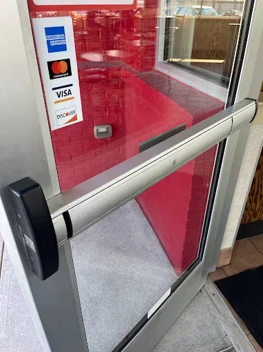 A glass door with a silver push-bar panic handle, displaying payment method decals for American Express, Mastercard, and Visa.