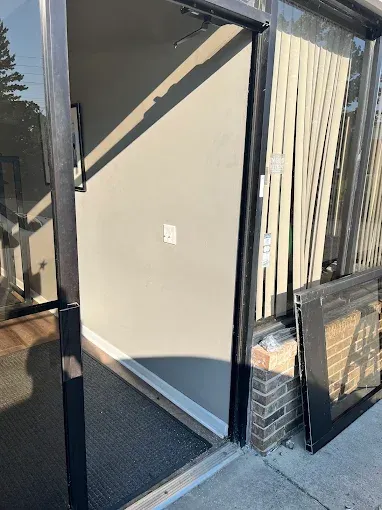 An open glass storefront door leading to a gray entryway with a floor mat and vertical blinds visible to the right.