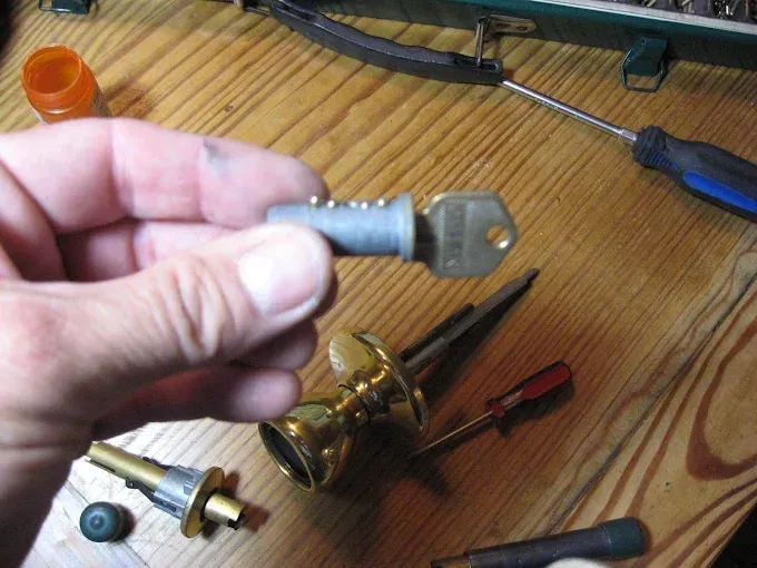 A hand holding a metal lock cylinder with a key inserted, next to a disassembled brass doorknob on a wooden table.