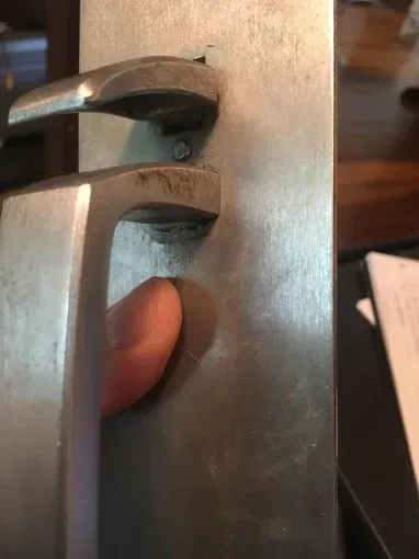 A finger presses on a metallic door handle assembly with a thumb turn lock mechanism on a rectangular backplate.