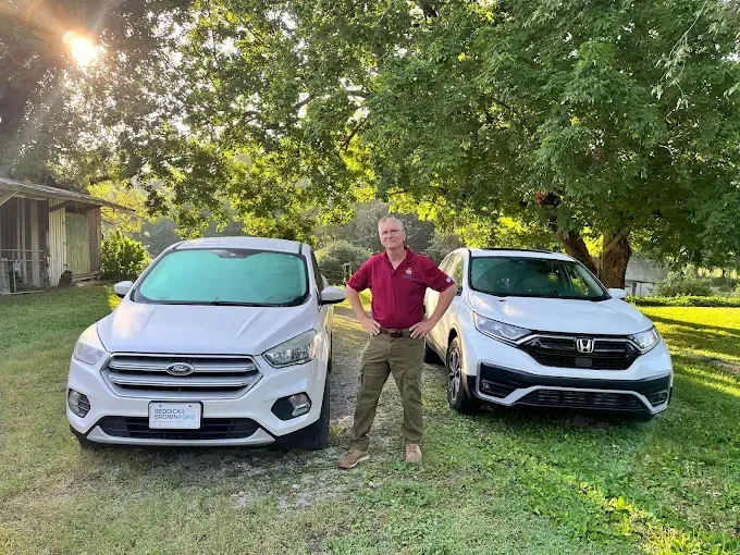 A person stands confidently with hands on hips between a white Ford Escape and a white Honda CR-V in a grassy yard.
