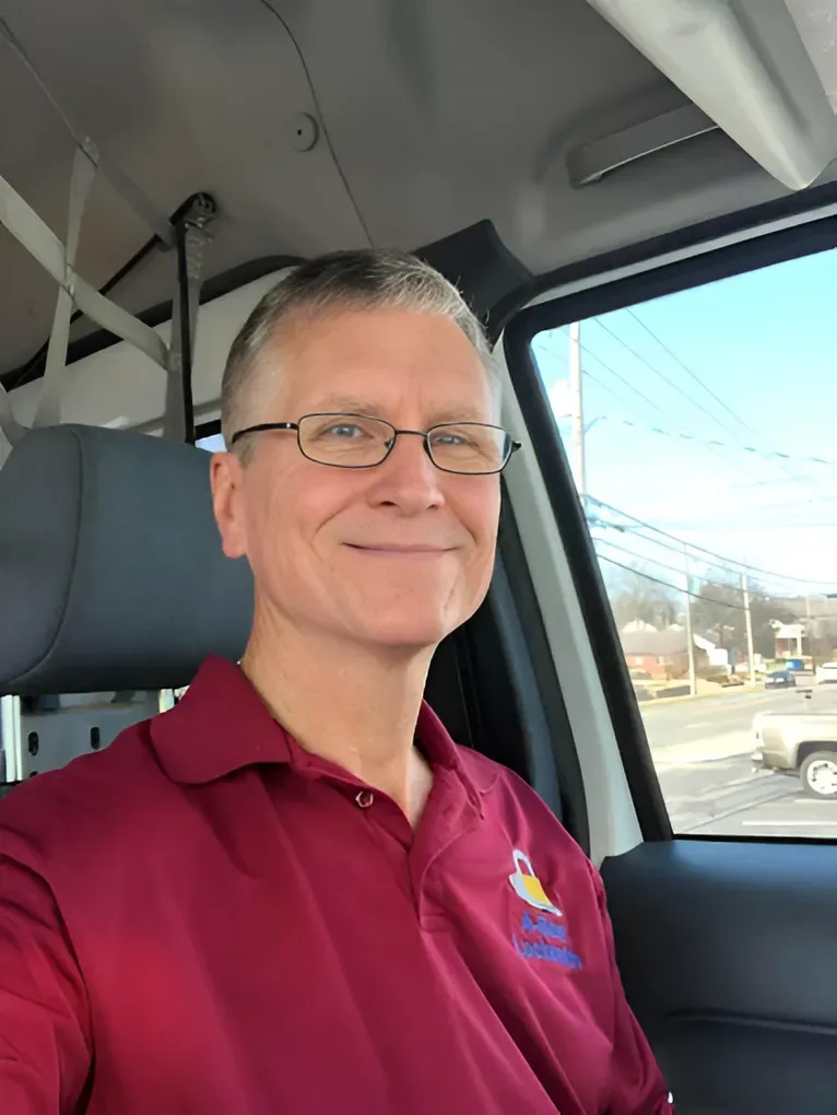 A person with glasses smiles while sitting in the driver's seat of a vehicle on a sunny day.