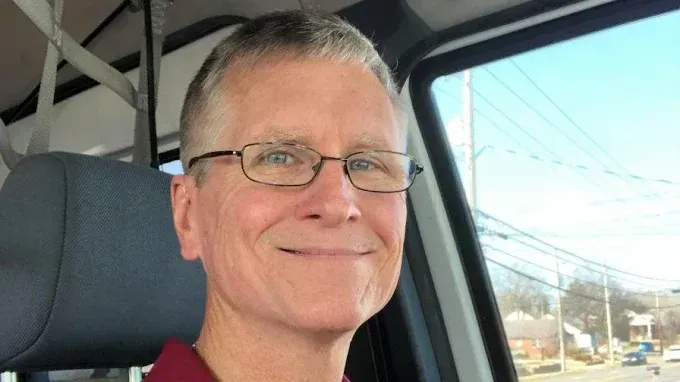 A smiling person wearing glasses, seated in the driver's seat of a vehicle with a view of a street visible through the window.