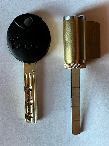 A brass-colored lock cylinder next to its corresponding dimple key with a black plastic head on a white background.