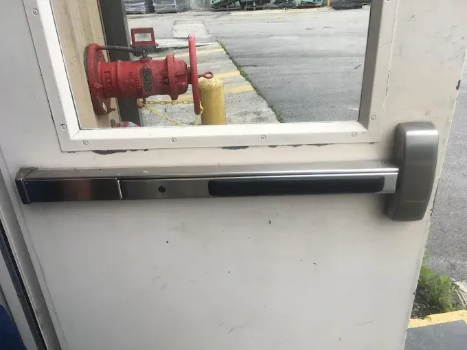 A close-up view of a metal crash bar exit device installed on a white door, with a red fire valve visible through the glass.