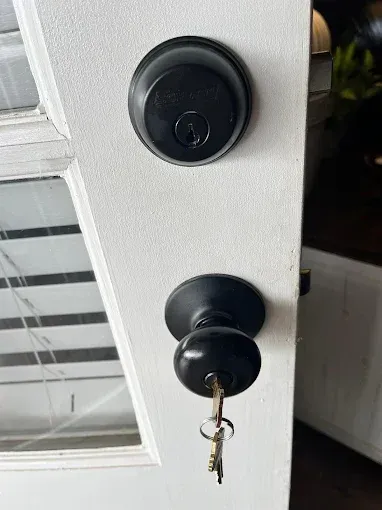 A matte black deadbolt and matching doorknob with a key inserted, mounted on a white door with a glass pane.