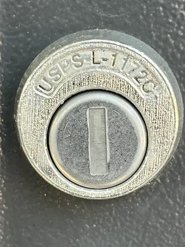 A silver, circular United States Postal Service mailbox lock labeled 
