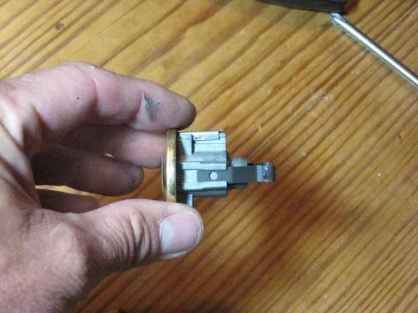 A hand holds a metallic door lock cylinder mechanism against a light wood background.