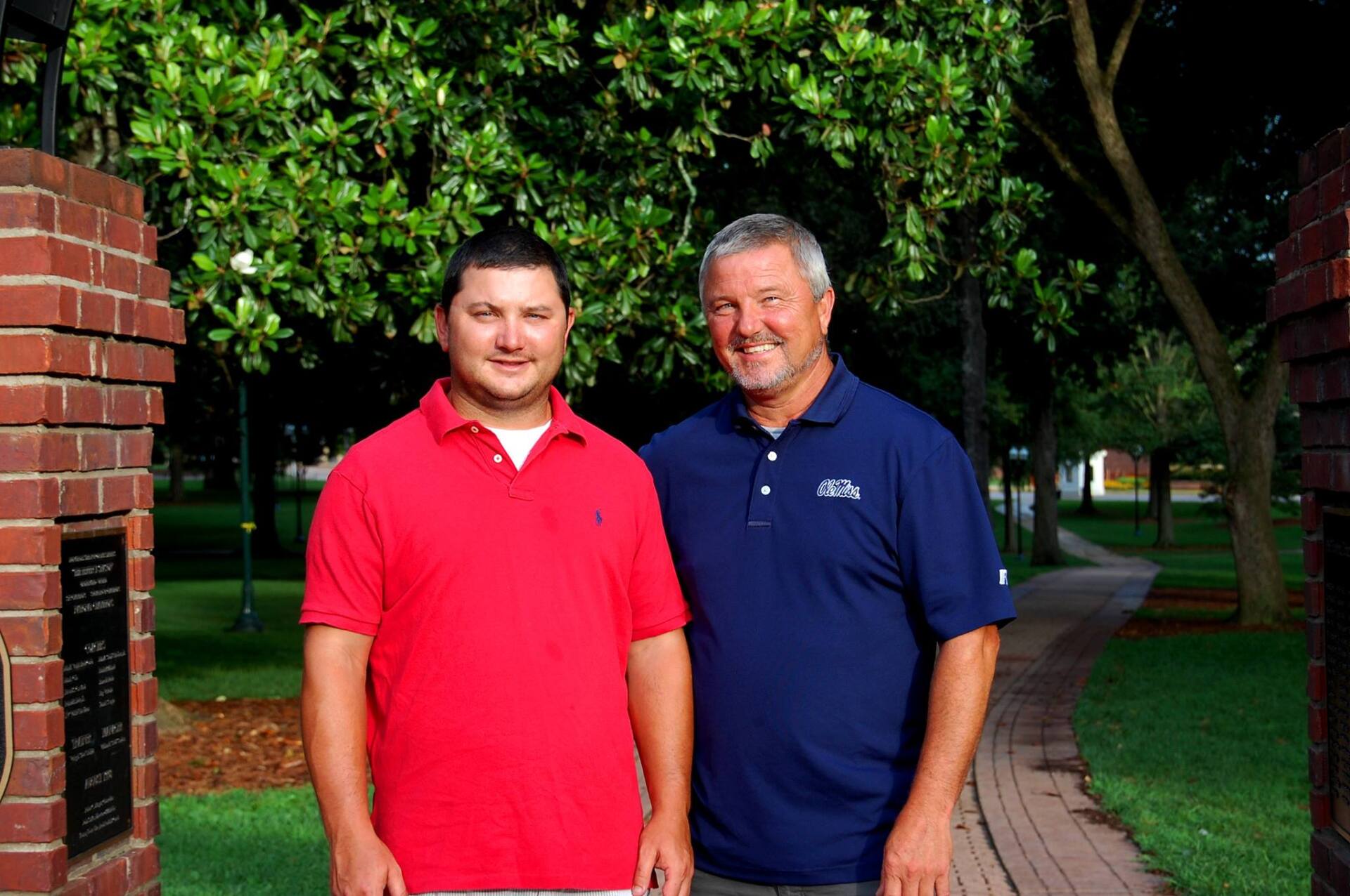 Two men stand side-by-side in front of brick pillars and trees, one in a red polo shirt and one in a navy blue polo shirt.