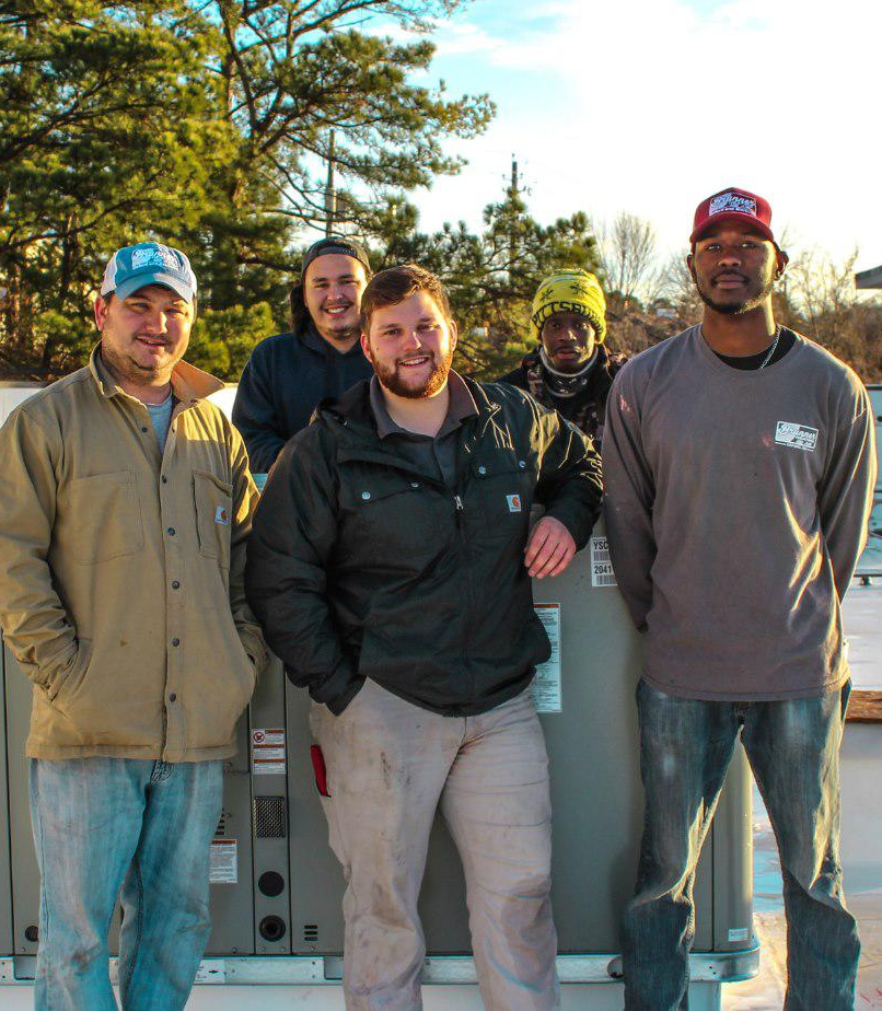 Five people stand together outdoors on a rooftop near an HVAC unit, smiling and posing for a group photo.
