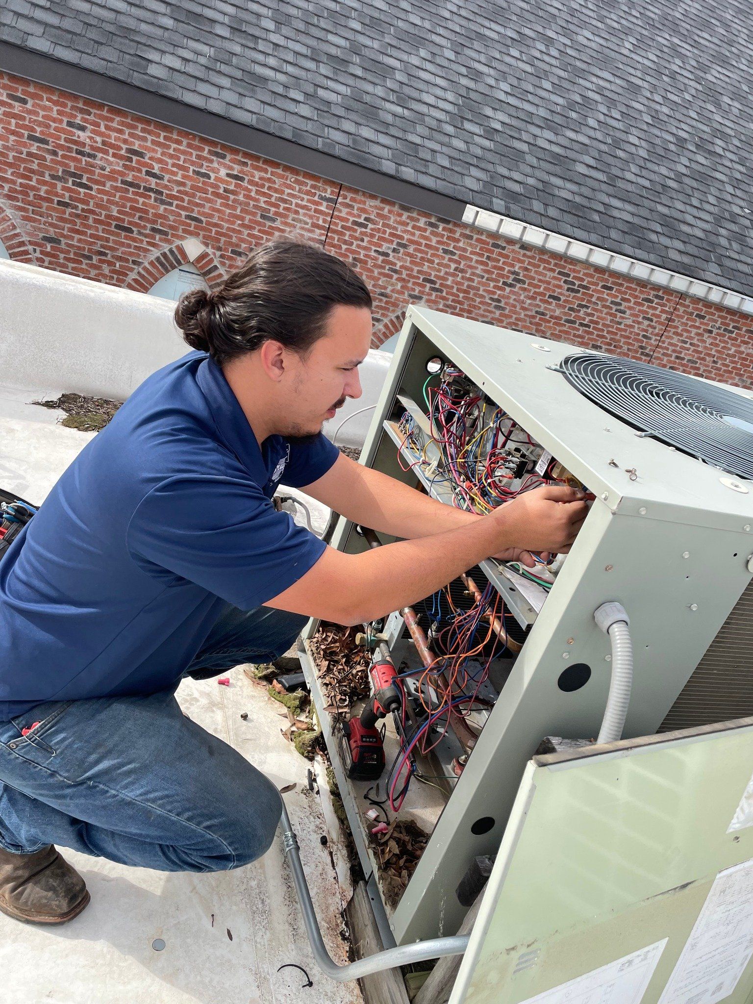 A technician in a blue polo shirt repairs the electrical components of an HVAC unit on a rooftop.