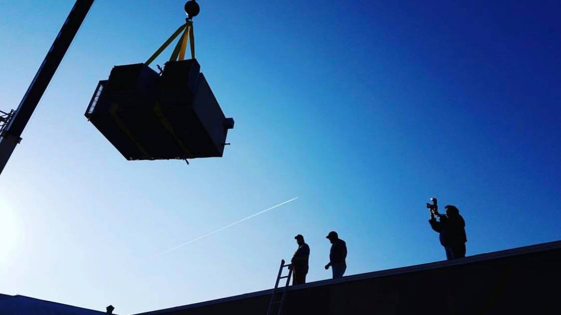 A crane lowers a large unit onto a building roof as three workers stand by and supervise the installation.