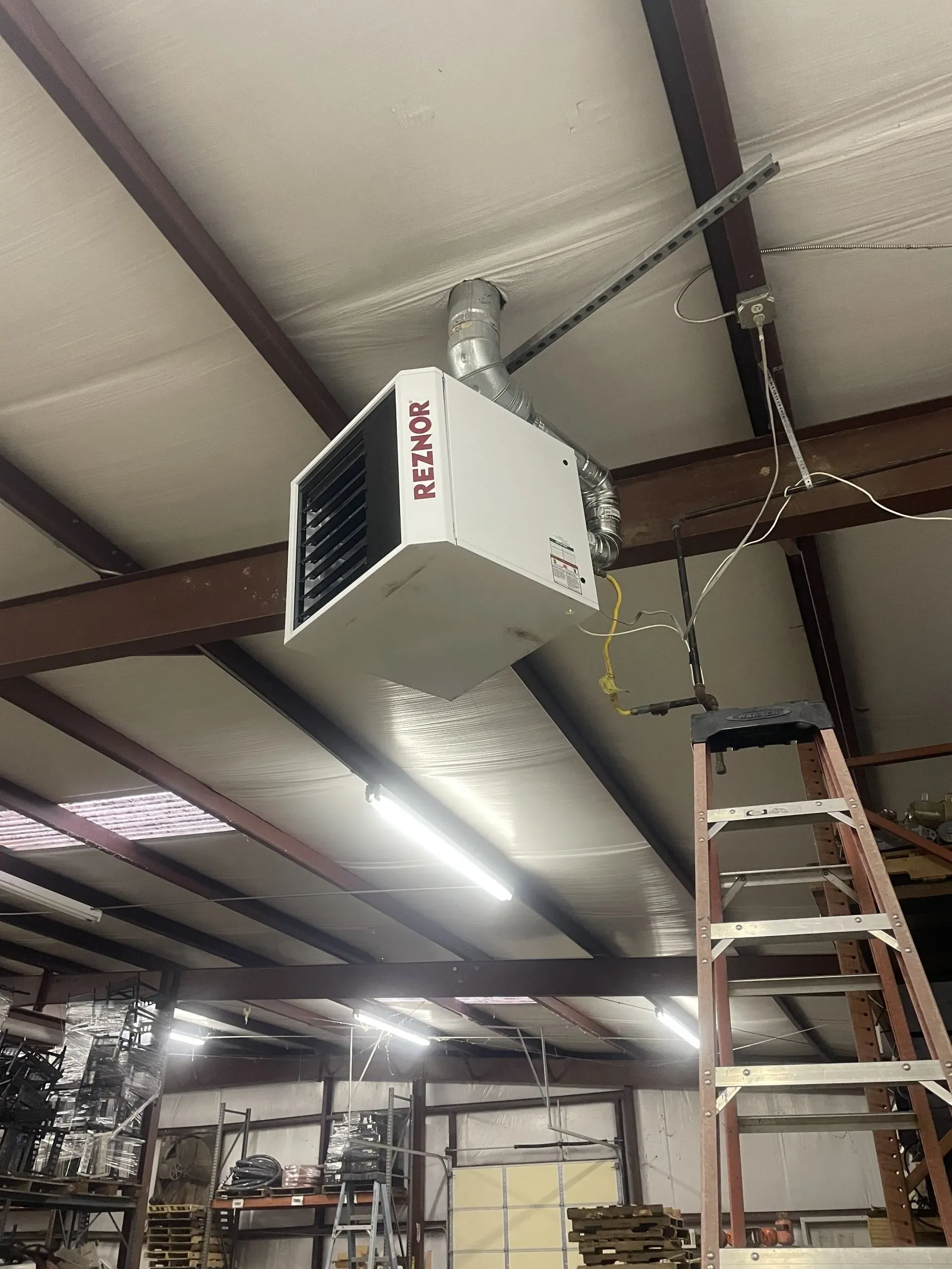 A white Reznor unit heater suspended from the ceiling of an industrial workshop next to a tall ladder.