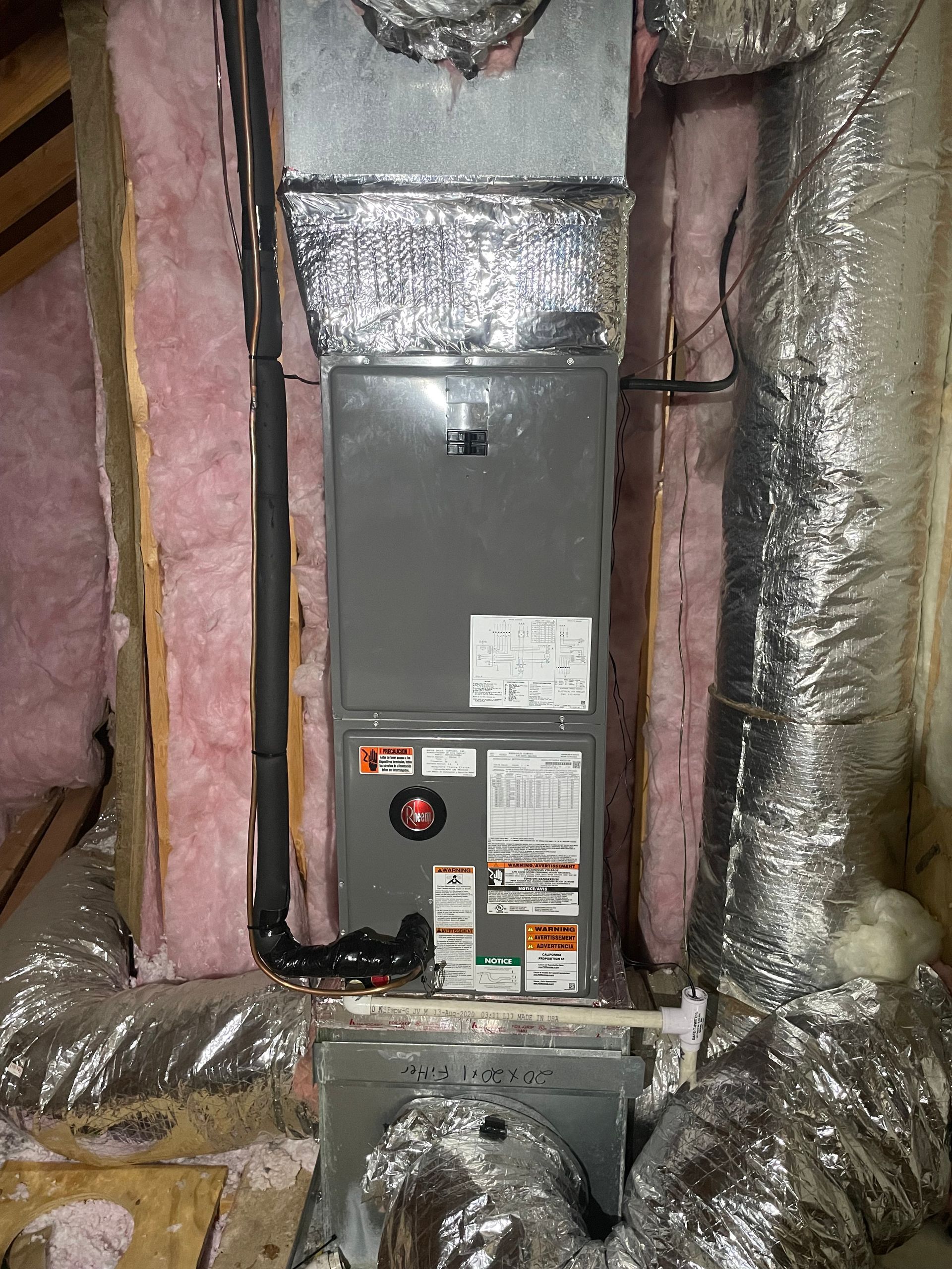 A gray HVAC air handler unit installed in an attic surrounded by pink fiberglass insulation and metallic ductwork.