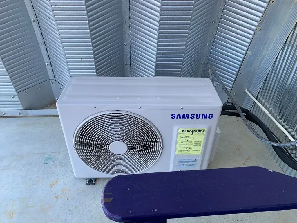A white Samsung air conditioning unit sits on a concrete floor in front of curved, corrugated metal panels.