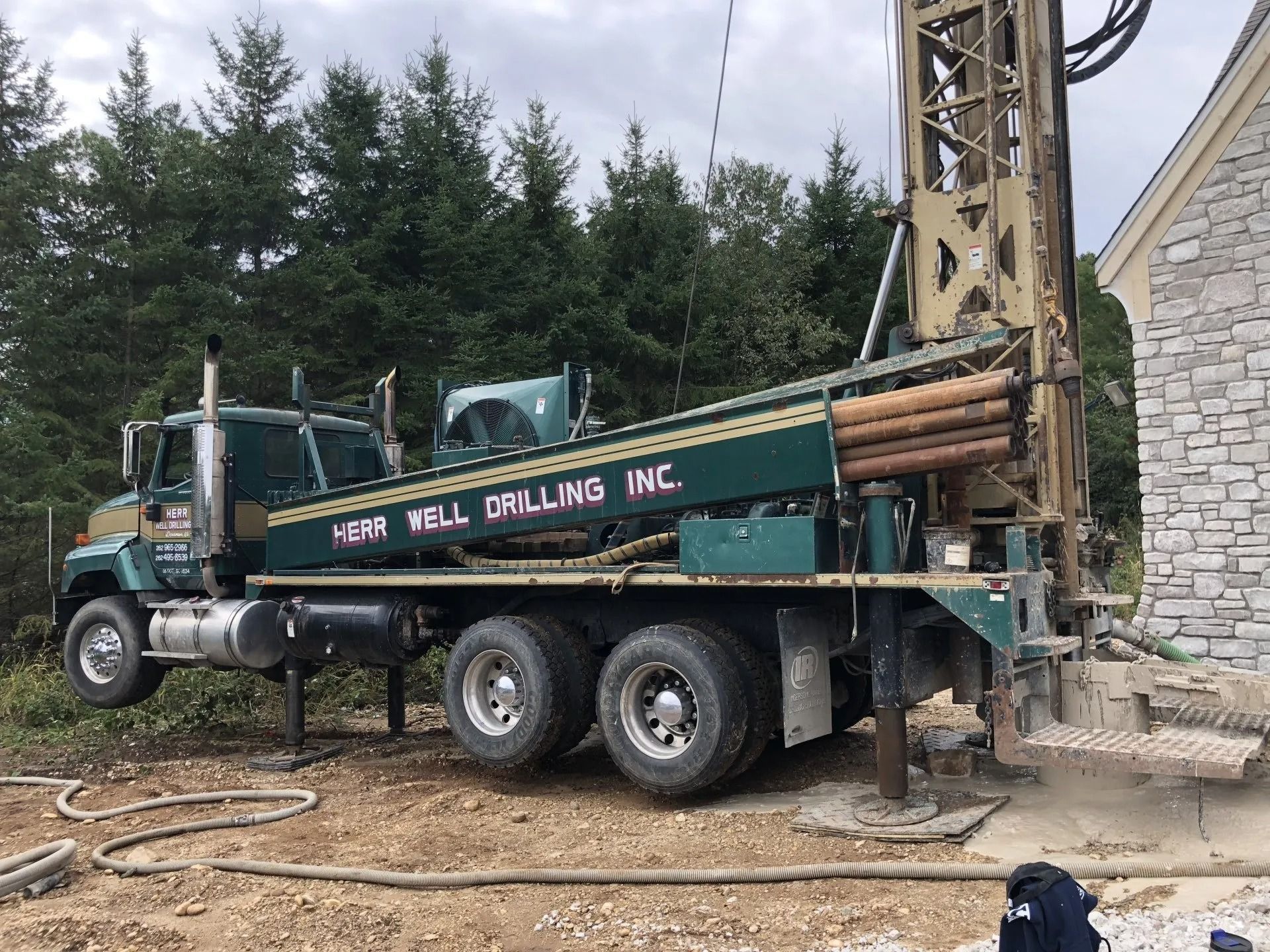A dark green Nor-Well Drilling Inc. truck is set up to drill a well next to a stone house in a wooded area.