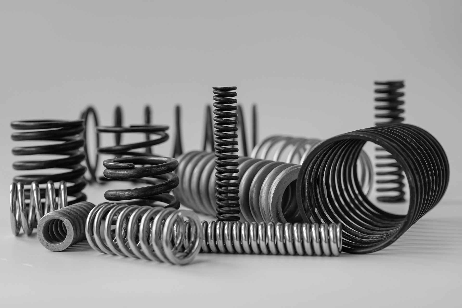 High-quality compression spring supplier parts, close-up on a bright factory floor. Assorted metal coil springs in black and silver arranged on a light gray surface.