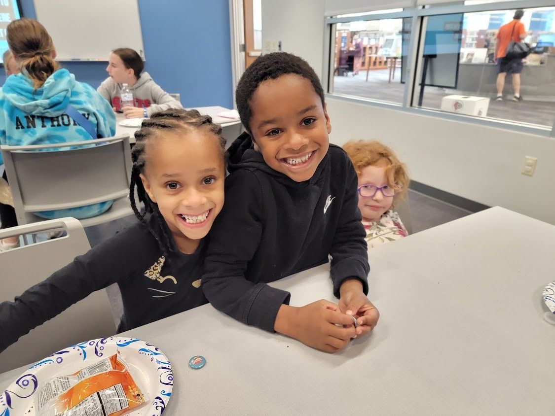 Two smiling children, one with braids, at a table. A third child peeks from behind them.