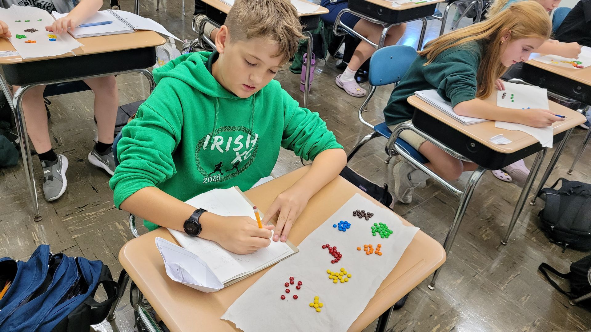 Students in a classroom working on a craft project at their desks. One boy wears a green sweatshirt.