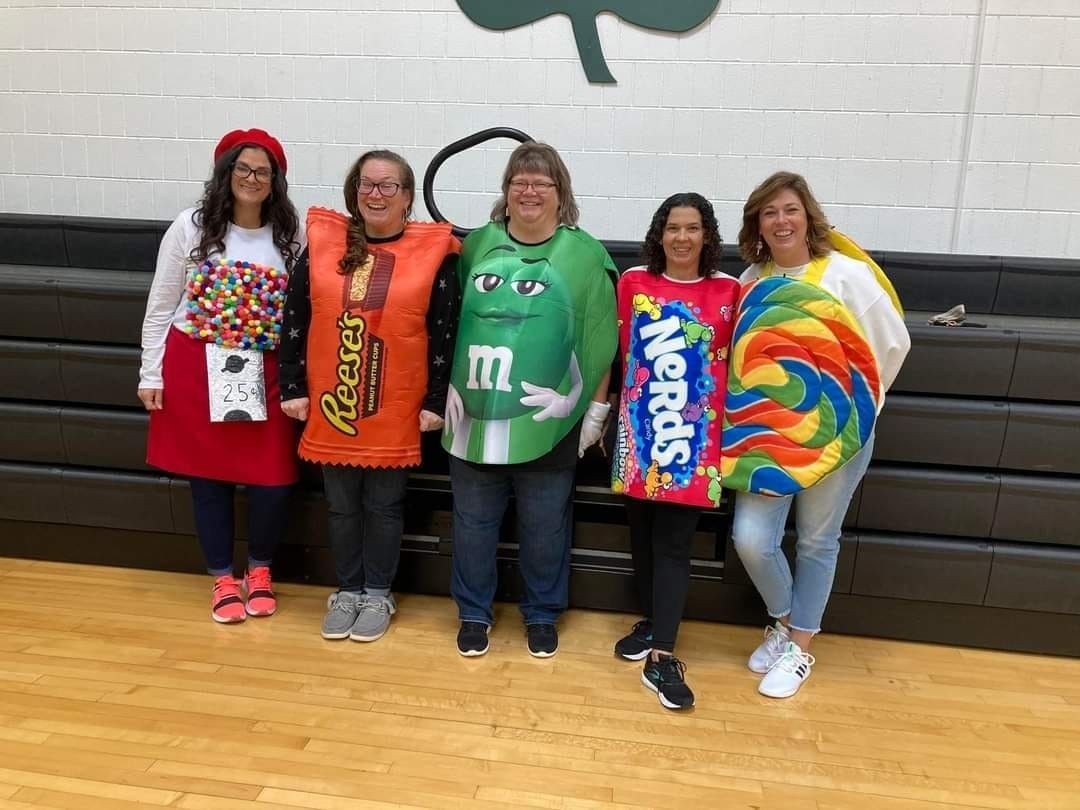 Five people dressed as candy in a gymnasium: M&M, Reese's, Nerds, and lollipop costumes.