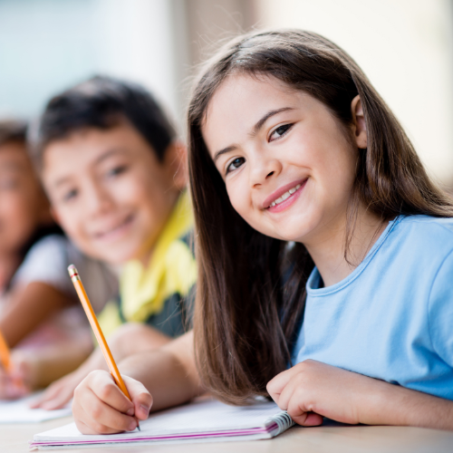 Girl with long brown hair smiles while writing in a notebook. Two other children in background.