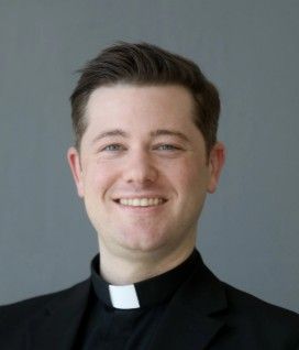 Priest with glasses and clerical collar smiling, against a gray backdrop.