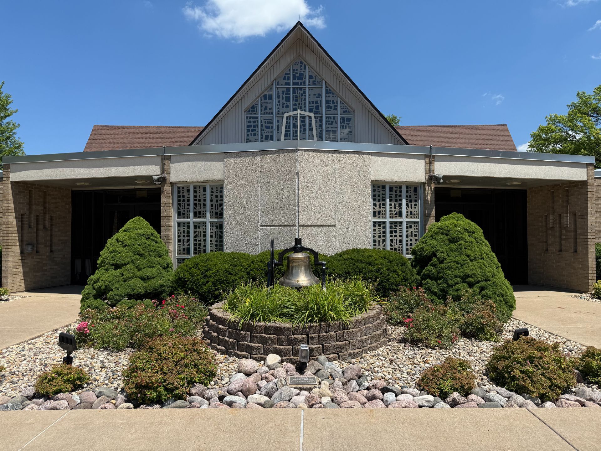 Church exterior with a bell, landscaping, and stained glass windows on a sunny day.