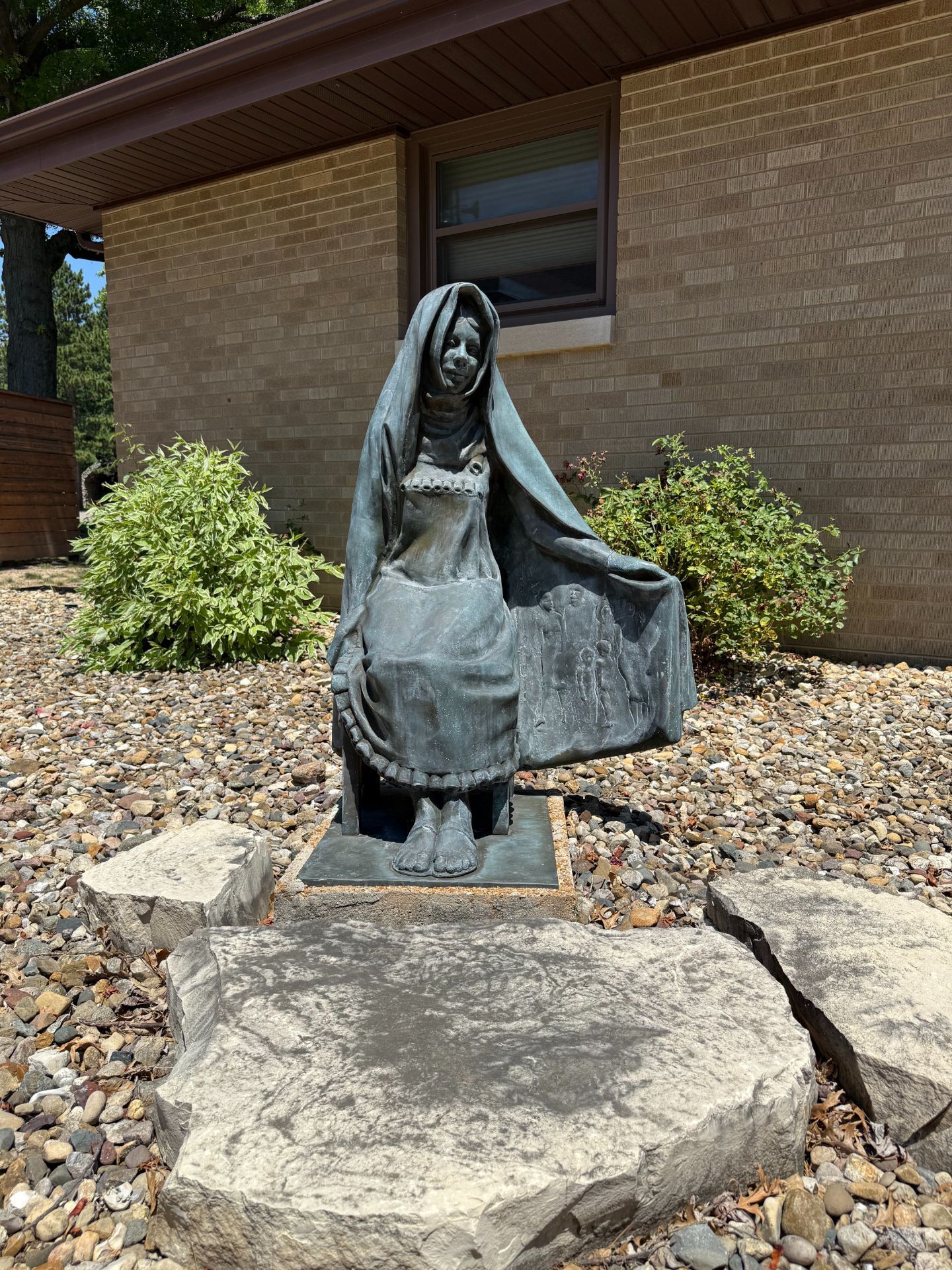 Bronze statue of a seated woman wearing a veil, in front of a brick building.