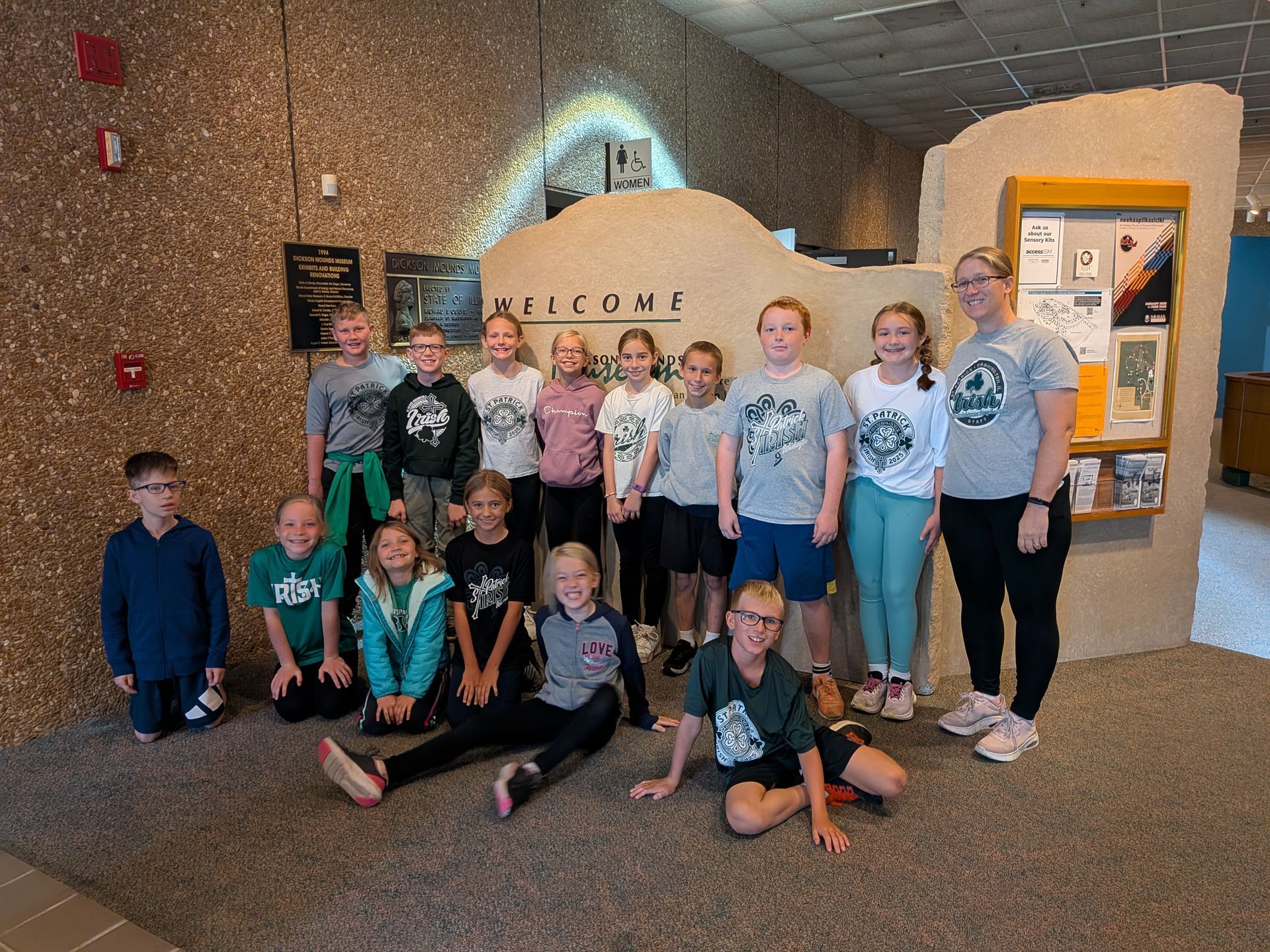 Group of kids in front of a welcome sign, inside a building. Some are kneeling, smiling.
