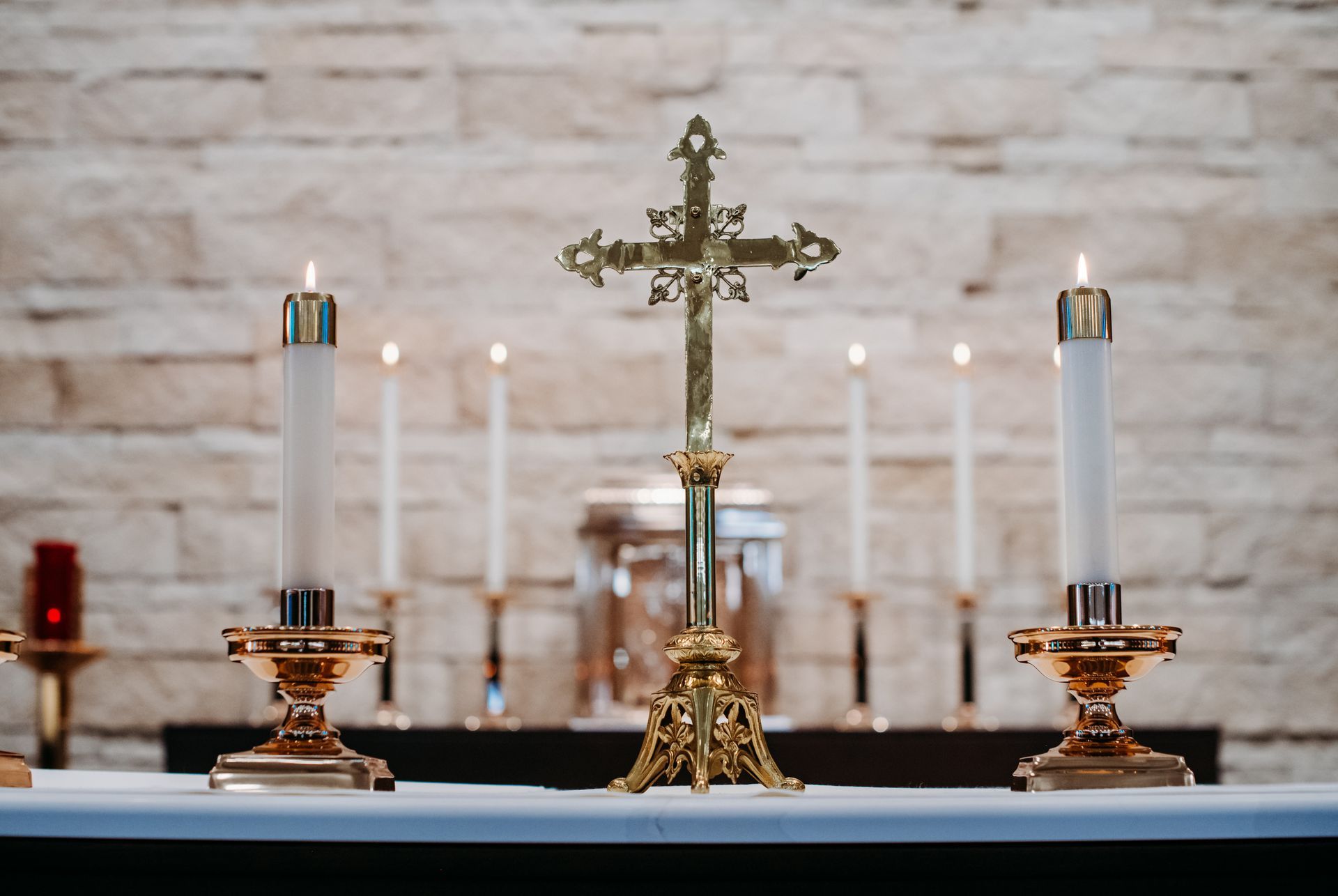 Gold cross flanked by lit candles on a white altar in front of a stone wall.