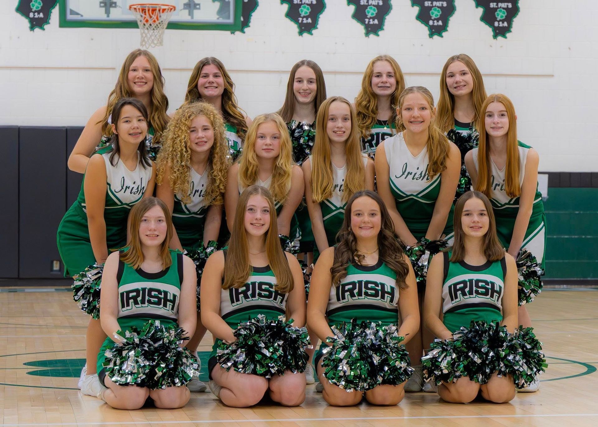 High school cheerleading squad in green and white uniforms pose with pom-poms on a basketball court.