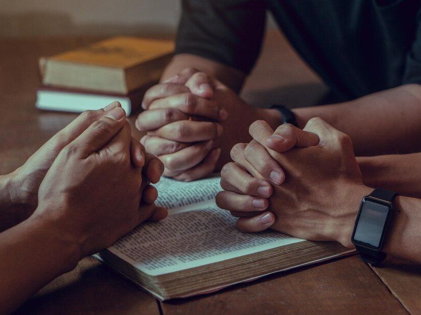 Hands clasped in prayer over an open book, presumably a Bible. Dark wood table, warm tones.
